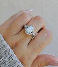 A sterling silver ring with a central lab-created opal surrounded by clear cubic zirconia stones, displayed on a person's hand.
